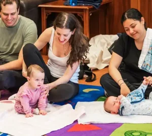 Three adults gather on a colorful mat with two babies; one baby crawls while the other rests on their back, sharing joyful moments.