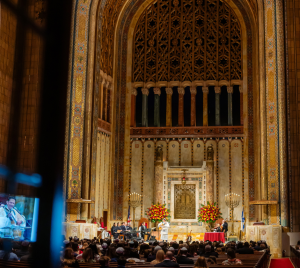 A large audience gathers for an event in the majestic sanctuary of Temple Emanu-El, surrounded by tall arches and elegant floral arrangements.