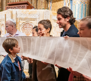 Congregants of all ages, including children, hold a Torah scroll with Hebrew text inside the beautifully adorned sanctuary at Temple Emanu-El.