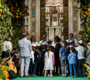 Children and adults gather around a beautifully decorated bimah with fruits, flowers, and greenery during a festive celebration at Temple Emanu-El.