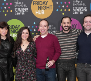 Seven people smiling, standing together in front of a colorful Friday Night Hub backdrop at Temple Emanu-El in New York City.