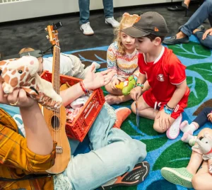A woman plays ukulele and smiles at children seated in a circle, each holding a stuffed animal, during a Temple Emanu-El program.