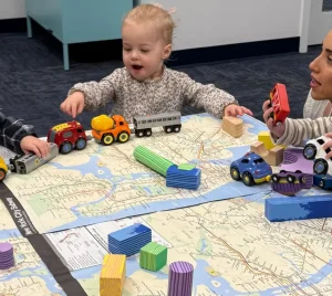 Three toddlers play with toy cars and blocks on a mat as an adult gently supervises, smiling warmly, in a synagogue classroom.