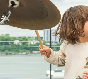 A young child joyfully plays a drum set outdoors near a waterfront railing during a community event at Temple Emanu-El.