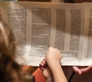 Children learning from an open Torah scroll with Hebrew text at Temple Emanu-El, guided by a caring adult.