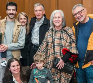 Seven adults and two young boys smile together in front of a wood-paneled wall at Temple Emanu-El in New York City.