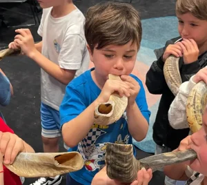 Children stand in a circle, joyfully blowing shofars together in a bright classroom at Temple Emanu-El.