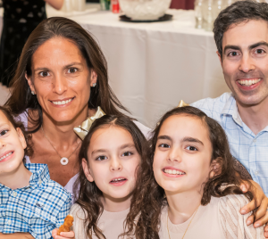A family of four, two adults and two children, share a warm smile together at a table inside Temple Emanu-El’s bright communal space.