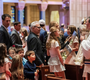 A rabbi welcomes children and adults holding prayer books in the sanctuary at Temple Emanu-El, with sunlight filtering through the ornate stained glass windows.