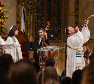 Four musicians dressed in white robes perform on stage at Temple Emanu-El; one musician sounds a shofar as the audience looks on.