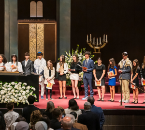 A group of young people stand on the bimah at Temple Emanu-El during a ceremony, proudly holding certificates.