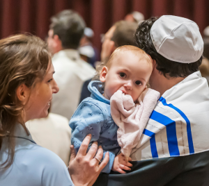 A baby in a blue outfit is held by an adult during a lively gathering at Temple Emanu-El, with community members present in the background.
