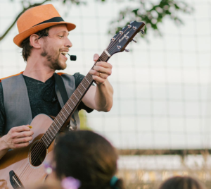 Man in orange hat playing guitar and singing outdoors at a Temple Emanu-El event, with attendees watching in the foreground.