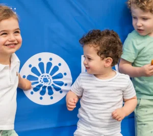 Three children smile in front of a blue banner at Temple Emanu-El, one holding a snack and water bottle.