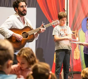 A man plays guitar on stage at Temple Emanu-El while children gather and interact in front of a vibrant backdrop.
