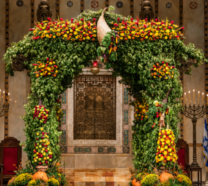 A spacious, beautifully decorated sukkah adorned with green leaves, yellow and orange flowers, and hanging fruit inside Temple Emanu-El in New York City.