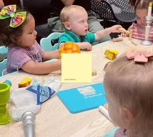 Six young children gather around a table playing with toys, guided by adults in a welcoming Temple Emanu-El classroom setting.