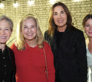 Four women smiling together at a Temple Emanu-El event, standing indoors in front of a brick wall adorned with lights.