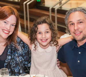 Three people smiling together at a table inside Temple Emanu-El, with a child in the middle warmly embracing two adults.