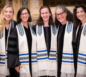 Five women in robes and white tallitot smile together inside Temple Emanu-El's sanctuary.