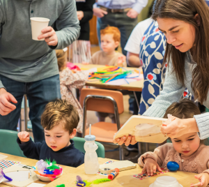 Adults and children creating crafts together at a busy table in a colorful, lively Temple Emanu-El classroom setting.