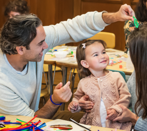 A family helps their toddler with a craft project at a colorful, busy classroom table in Temple Emanu-El’s religious school.