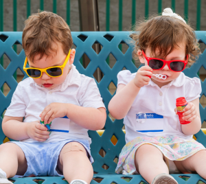 Two young children wearing sunglasses sit together on a bench; one blows bubbles while the other examines a bubble wand.