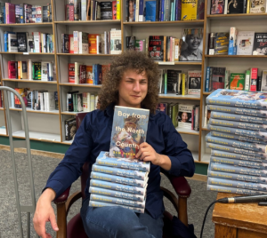 Individual seated in a bookstore holding a book, with additional copies neatly stacked on a nearby table, representing an engaging moment of learning and community.