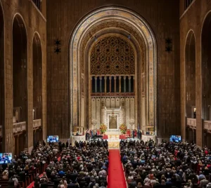 A large congregation gathers inside Temple Emanu-El’s grand sanctuary, with soaring arches and a red carpet down the central aisle.