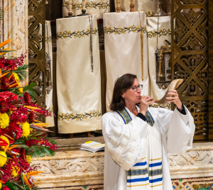 A person in a white robe blows a shofar inside Temple Emanu-El, with vibrant floral arrangements visible on the left.