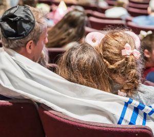 A man wearing a tallit embraces two children while seated among a large gathering in the Temple Emanu-El sanctuary.