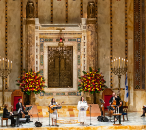 Musicians and vocalists perform in the grand, ornate sanctuary of Temple Emanu-El, with large floral arrangements and decorative elements displayed.