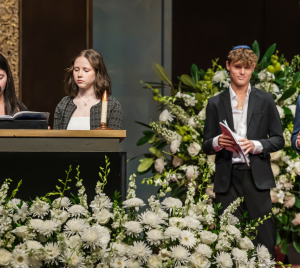 Four young people stand by a podium decorated with flowers in the sanctuary of Temple Emanu-El; two are speaking while the other two hold prayer books and listen attentively.