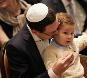 An adult wearing a kippah warmly embraces a young boy amid a gathering, both dressed in formal attire.