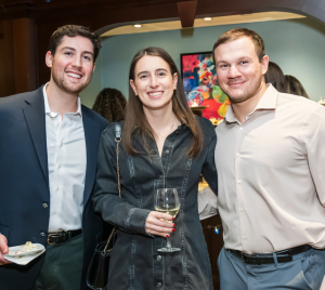 Three people smiling at a Temple Emanu-El gathering, one holding a plate, another with a glass, with others mingling in the background.