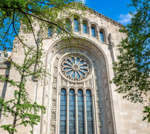 Large arched stained-glass window on the historic façade of Temple Emanu-El, framed by green tree branches with a backdrop of blue sky.