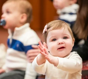Smiling baby claps hands while sitting with children and adults during a community gathering at Temple Emanu-El.