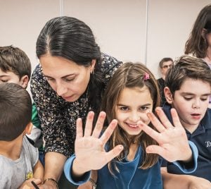 Smiling girl displaying her painted hands in a bright classroom at Temple Emanu-El, with children and adults gathered around her.