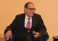 Distinguished older man in a suit and glasses smiles while holding a microphone at Temple Emanu-El, with water on a table nearby.