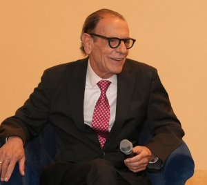 Distinguished older man in a suit and glasses smiles while holding a microphone at Temple Emanu-El, with water on a table nearby.
