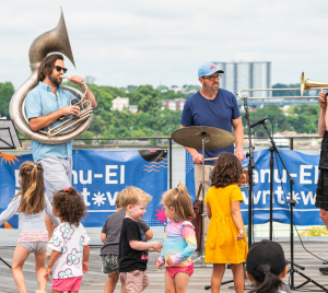Three musicians play on an outdoor stage at Temple Emanu-El as children dance and enjoy the sunny day in front of them.
